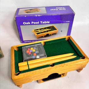 May include: A miniature wooden pool table with a green felt surface, two light-colored wooden cues, and a clear plastic case containing small colorful billiard balls. The set is displayed in front of a purple box labeled Executive Oak Pool Table.