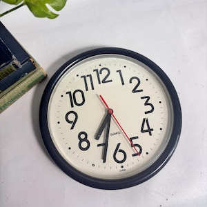 May include: A round wall clock with a black frame and an off-white face. The clock features large black Arabic numerals from 1 to 12, black hour and minute hands, and a thin red second hand. The word QUARTZ is printed near the center, and CHINA is at the bottom.