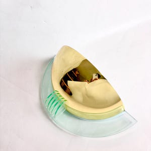 Può includere: Un'applique semicircolare con un paralume curvo in metallo giallo pallido e una superficie interna riflettente. La base è realizzata in vetro trasparente azzurro con una serie di quattro alette verticali in vetro verde chiaro su un lato.