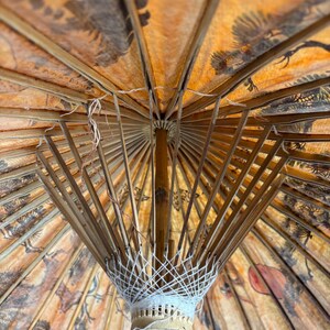 Puede incluir: Un parasol antiguo con estructura de bamb&uacute; y tela naranja estampada. La tela presenta ilustraciones en negro y marr&oacute;n de p&aacute;jaros y &aacute;rboles. La estructura interior del parasol es visible, mostrando su intrincada construcci&oacute;n.