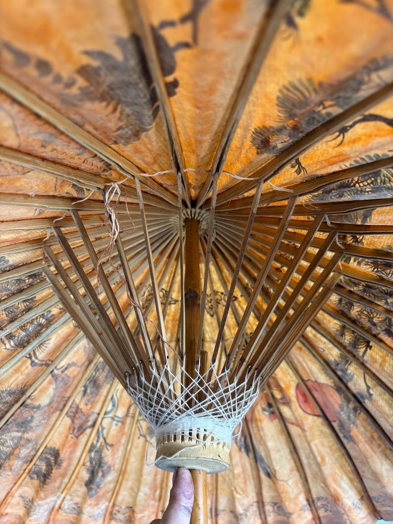 Puede incluir: Un parasol antiguo con estructura de bamb&uacute; y tela naranja estampada. La tela presenta ilustraciones en negro y marr&oacute;n de p&aacute;jaros y &aacute;rboles. La estructura interior del parasol es visible, mostrando su intrincada construcci&oacute;n.