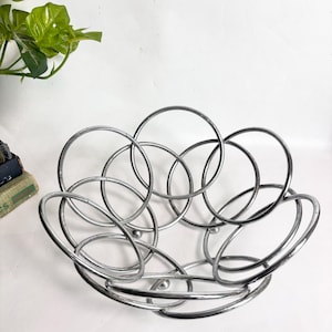 Op de afbeelding: Een zilverkleurige metalen fruitschaal opgebouwd uit een reeks onderling verbonden cirkelvormige ringen. De schaal heeft een afgerond ontwerp met een open frame, een gepolijste afwerking en kleine bolvormige voetjes aan de onderkant.
