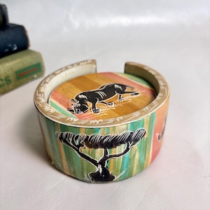 May include: A round wooden coaster holder featuring a painted black acacia tree on a green and orange striped background. The top rim is decorated with white etched patterns, and the interior contains a coaster with a black rhinoceros illustration.