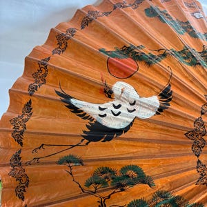 Puede incluir: Un abanico de papel naranja con una grulla pintada en vuelo, un sol rojo y &aacute;rboles verdes. El abanico tiene un borde negro decorativo. La grulla es blanca con puntas de alas negras. El abanico est&aacute; abierto y desplegado.