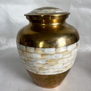 May include: A gold-colored urn with a lid, featuring a band of white and cream-colored mosaic tiles around the middle. The urn has a rounded shape and a polished finish, suitable for memorial purposes.