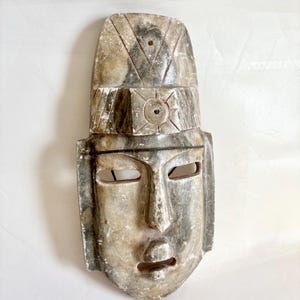 May include: A carved stone mask with a rectangular shape and a detailed headdress. The mask has eye and mouth openings, and the stone has a mottled gray and beige appearance with red accent lines. The mask is likely a decorative or ceremonial item.