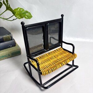 Peut inclure: Une petite chaise à bascule en métal noir avec une assise en osier jaune et deux cadres photo recouverts de verre. La chaise a un design décoratif avec des fleurettes arrondies sur le dessus. Le design général rappelle un style vintage ou rustique.