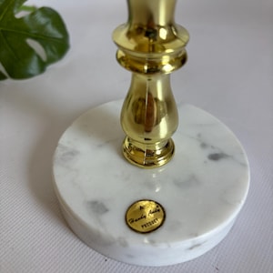 May include: A gold-colored metal candlestick holder with a white marble base. The base is round with gray veining. The candlestick has a decorative design and the words "Handy Andy Product" are on a gold sticker on the base.