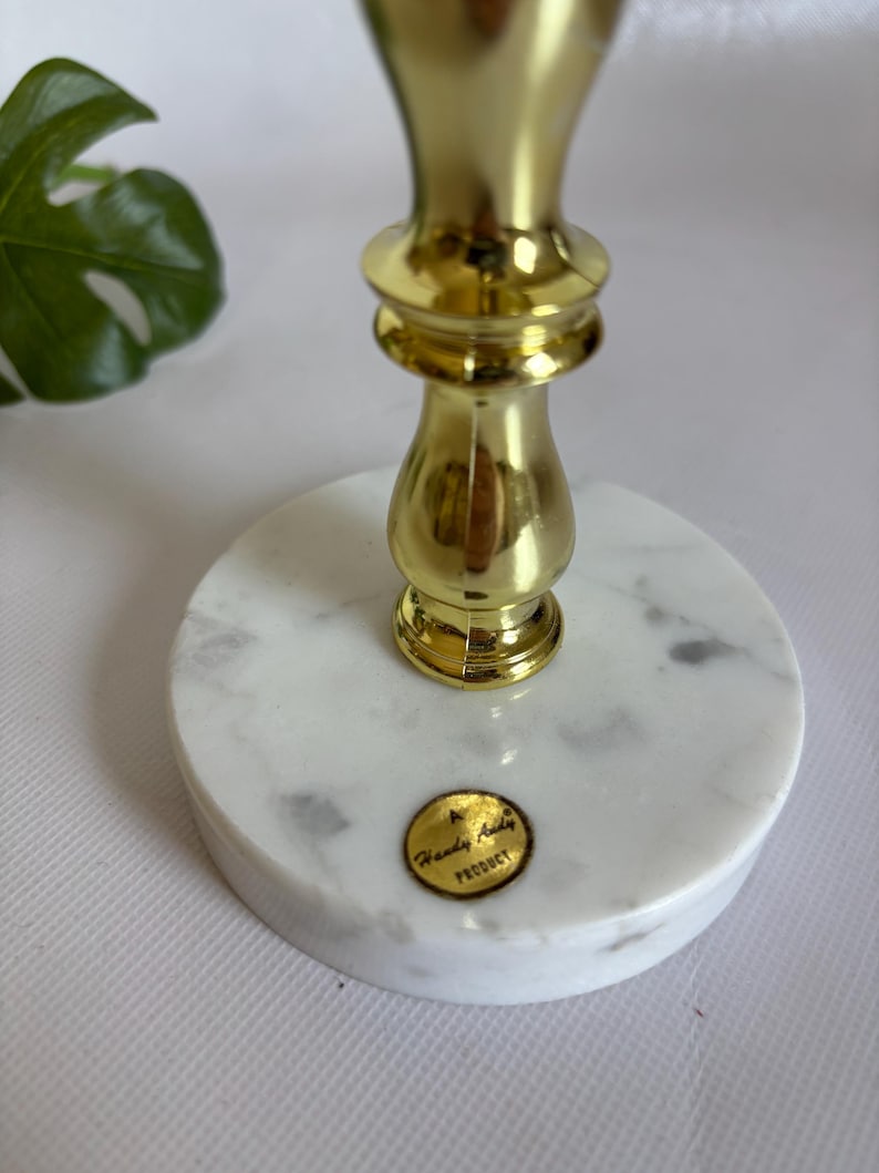May include: A gold-colored metal candlestick holder with a white marble base. The base is round with gray veining. The candlestick has a decorative design and the words "Handy Andy Product" are on a gold sticker on the base.