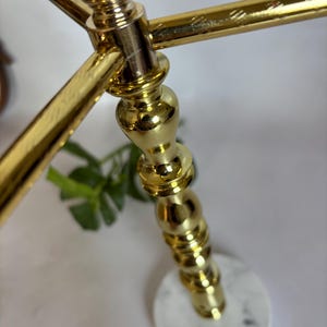 May include: A gold-colored metal decorative stand with a round, white marble base. The stand has a detailed, ornate design with a crossbar at the top. The base has a small, gold-colored emblem.
