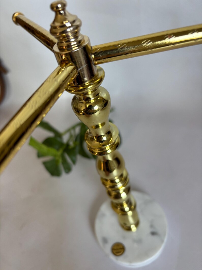 May include: A gold-colored metal decorative stand with a round, white marble base. The stand has a detailed, ornate design with a crossbar at the top. The base has a small, gold-colored emblem.
