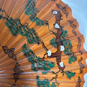 Puede incluir: Un abanico decorativo naranja con un dise&ntilde;o pintado a mano. El abanico presenta un &aacute;rbol central con follaje verde y detalles blancos, rodeado de patrones florales negros y verdes. El dise&ntilde;o del abanico recuerda al arte asi&aacute;tico tradicional.