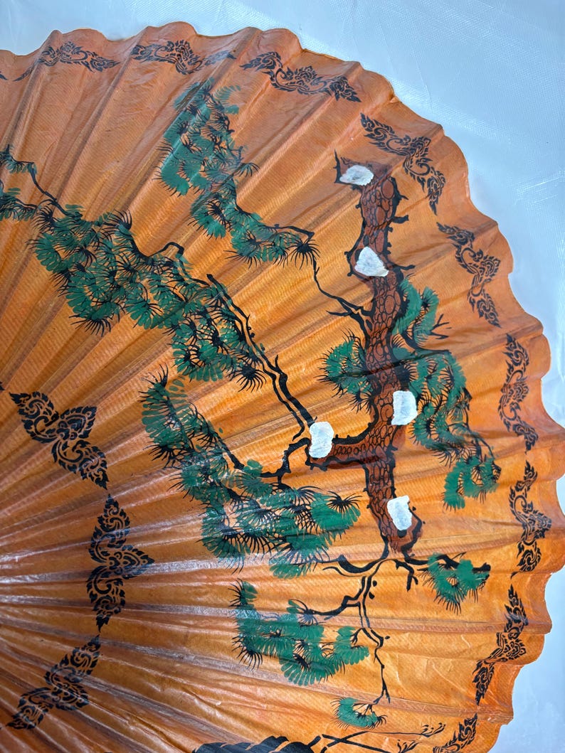 Puede incluir: Un abanico decorativo naranja con un dise&ntilde;o pintado a mano. El abanico presenta un &aacute;rbol central con follaje verde y detalles blancos, rodeado de patrones florales negros y verdes. El dise&ntilde;o del abanico recuerda al arte asi&aacute;tico tradicional.