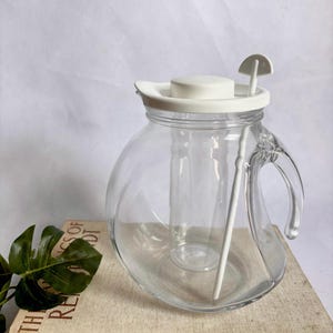 May include: A clear glass pitcher with a rounded body and a curved handle. It features a white plastic lid with a central circular knob and an attached white stirring rod that extends into the pitcher. The design includes an internal glass cylinder.