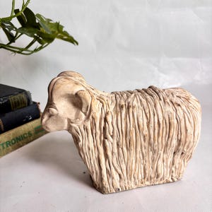 May include: A cream-colored, textured figurine of a ram with curled horns and a shaggy, vertical-lined coat. The sculpture features a weathered, off-white finish with subtle brown shading in the grooves of the carved wool texture.