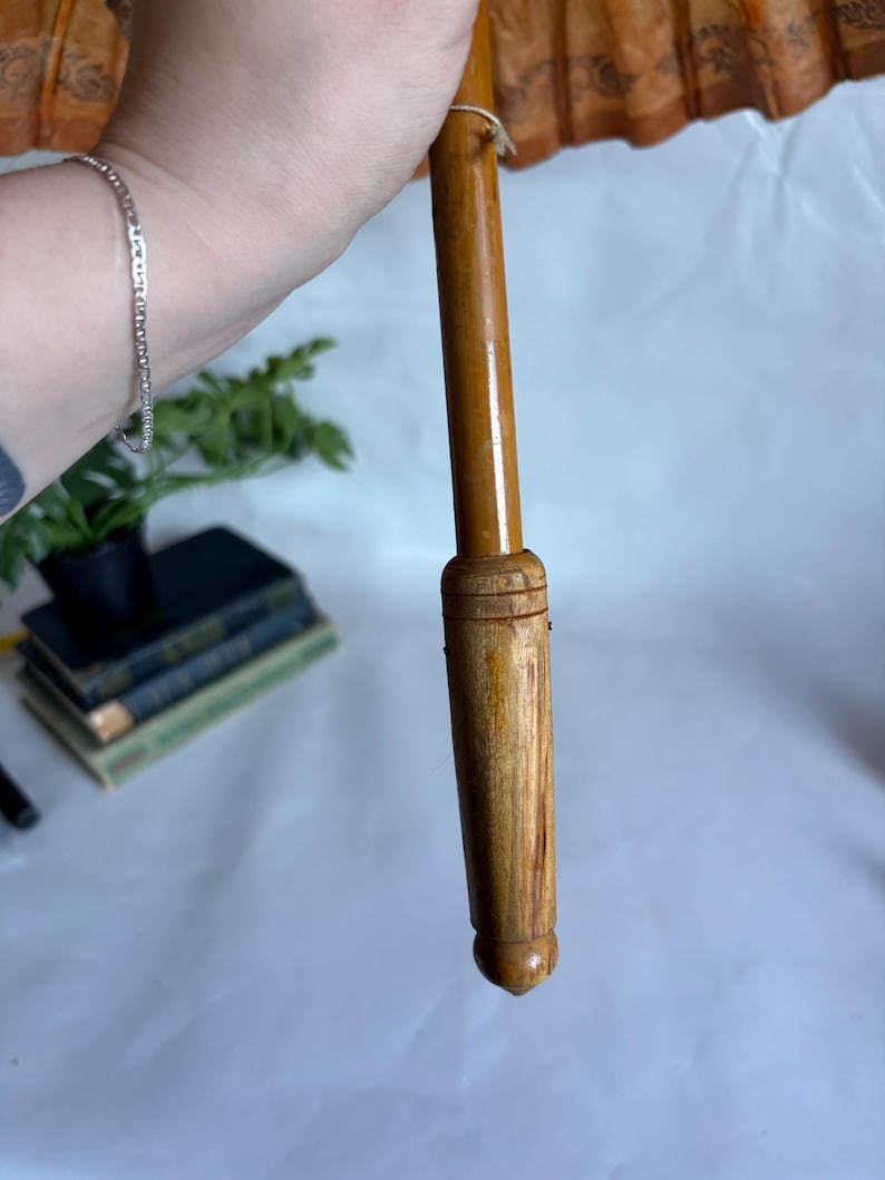 Puede incluir: Un parasol de estilo vintage con un mango de madera y una capota naranja decorativa. El mango es de color marr&oacute;n claro y la capota tiene un patr&oacute;n detallado. El parasol est&aacute; sostenido por una persona.