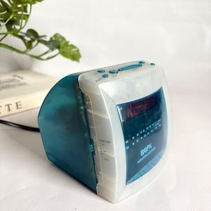 May include: A white and teal digital alarm clock with a curved design. The clock has a digital display, buttons for alarm and sleep functions, and an AM/FM radio. The brand name "GPX" is visible on the front.