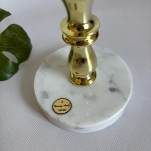 May include: A gold-colored metal candlestick with a marble base. The candlestick has a decorative design and a round marble base. The base has a sticker that says "A Handy Andy Product".