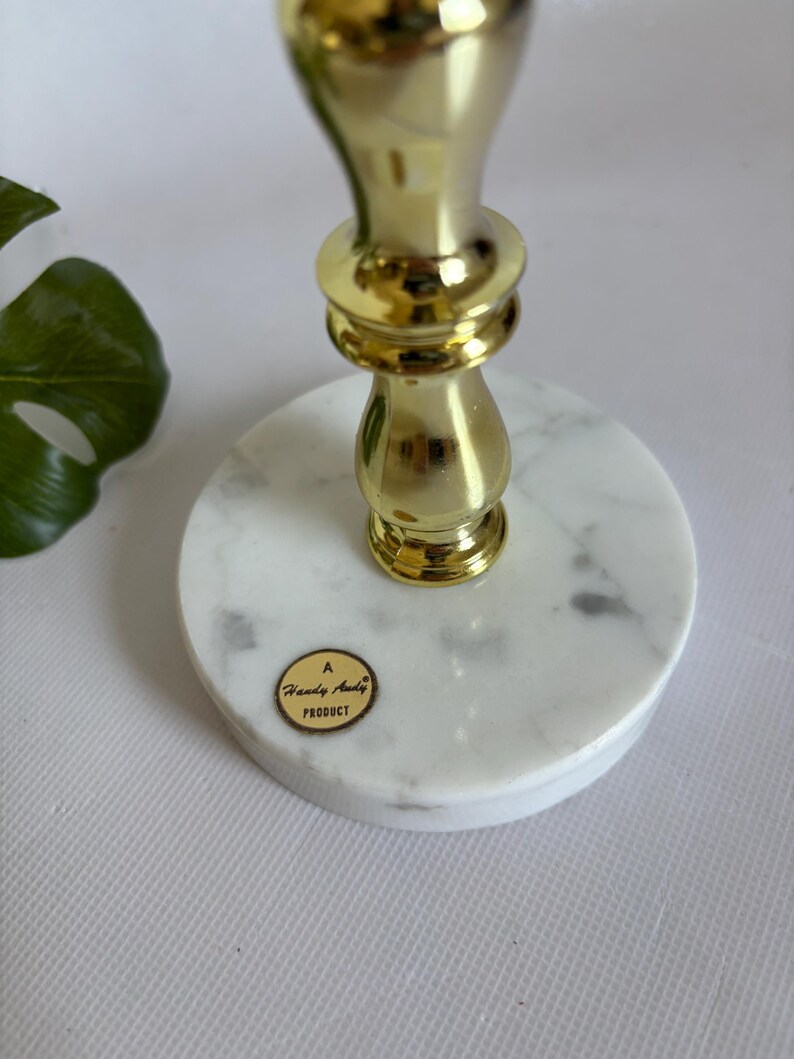 May include: A gold-colored metal candlestick with a marble base. The candlestick has a decorative design and a round marble base. The base has a sticker that says "A Handy Andy Product".