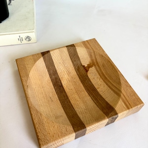 May include: Square wooden bowl with a circular indentation. The bowl features alternating light and dark brown stripes. The wood grain is visible, and the bowl appears to be handmade. The bowl is on a white surface.