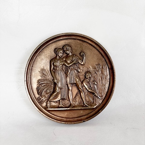 Op de afbeelding: Een bronzen, cirkelvormig reliëfbeeldhouwwerk met klassieke figuren. Het reliëf toont twee figuren die elkaar omarmen, met een derde figuur die knielt. Tarwearen zijn zichtbaar op de achtergrond. Het beeldhouwwerk heeft een gedetailleerd, antiek uiterlijk.