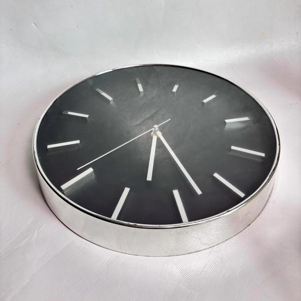 May include: A round wall clock featuring a dark charcoal gray face with a smooth, matte finish. The clock has a polished silver-tone metal frame and thin, silver-tone hour and minute hands. Twelve silver-tone rectangular markers indicate the hours.