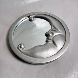 May include: A silver-colored, round mirror with a crescent moon design. The moon has a face sculpted into it, with a nose, closed eye, and a smiling mouth. The mirror is surrounded by a silver frame with decorative swirls.