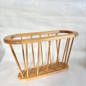 Puede incluir: Un revistero de madera de color claro con una parte superior e inferior ovaladas. Varillas verticales de madera conectan la parte superior e inferior, creando un diseño de listones. El estante está hecho de madera de color claro, con un acabado natural.