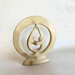 May include: A beige stone sculpture depicting two figures within a circular frame. The abstract design features a teardrop shape with the figures' forms. The sculpture has a smooth finish and a rounded base, suitable for display.