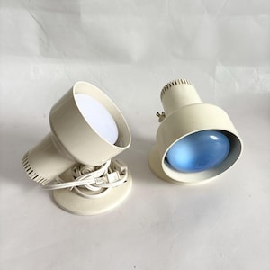 May include: Two off-white adjustable lamps with circular bases and cylindrical heads. One lamp features a bright white bulb, while the other contains a blue-tinted bulb. Each lamp includes a white power cord with a plug and a small toggle switch.