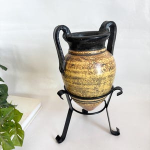 May include: A vintage-style ceramic vase with a black rim and handles, set on a black metal stand. The vase has a yellow and brown textured surface, evoking an aged aesthetic. The stand is approximately 6 inches tall.