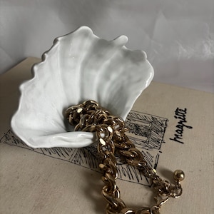 Puede incluir: Una pulsera de cadena dorada descansa en un plato de cerámica blanco con forma de concha. El plato está sobre un libro antiguo con el nombre "magritte" visible. La pulsera presenta eslabones grandes e interconectados y una pequeña bola en el cierre.