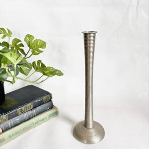 Op de afbeelding: Een hoge, zilverkleurige metalen kandelaar met een geribbeld ontwerp. De basis is cirkelvormig en de steel is lang en slank. De kandelaar wordt getoond met een stapel vintage boeken en een groene plant.