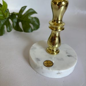 May include: A gold-colored metal candlestick with a marble base. The candlestick has a decorative, ornate design. The base is round, white, and gray-veined. A small, round sticker with text is affixed to the base.