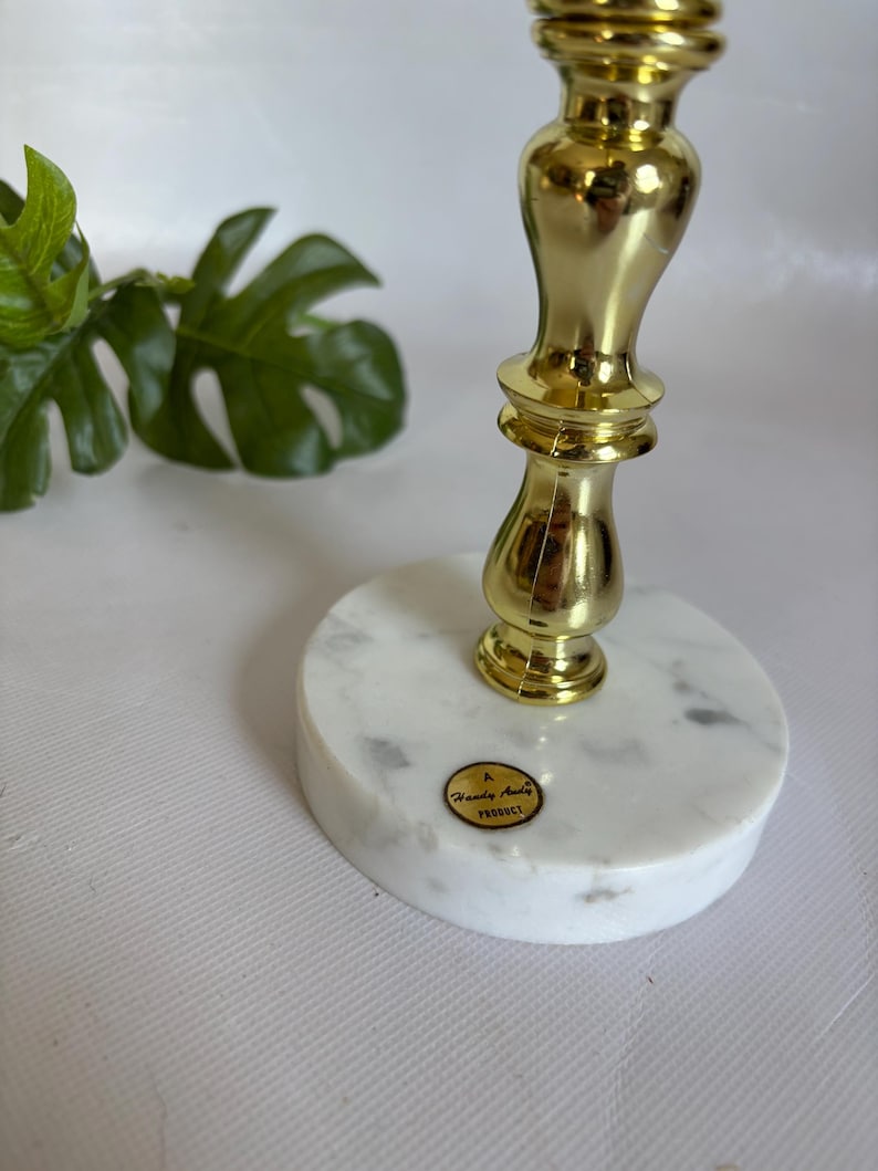 May include: A gold-colored metal candlestick with a marble base. The candlestick has a decorative, ornate design. The base is round, white, and gray-veined. A small, round sticker with text is affixed to the base.