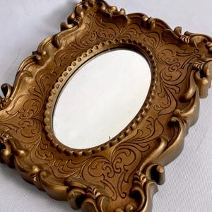 Puede incluir: Espejo ovalado dorado con un marco ornamentado con un patr&oacute;n de relieve arremolinado. El marco tiene un borde interior con cuentas. El dise&ntilde;o es vintage y elegante, adecuado para la decoraci&oacute;n del hogar. Medidas: aprox. 15 cm.