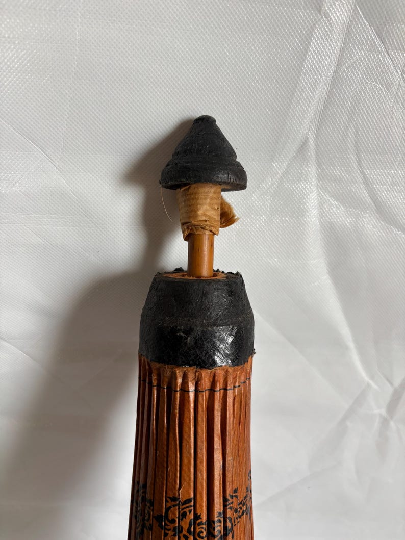 Puede incluir: Un peque&ntilde;o parasol decorativo con una parte superior negra y un cuerpo plisado marr&oacute;n. El mango es una clavija de madera fina envuelta en papel beige. El cuerpo del parasol tiene un dise&ntilde;o floral negro.