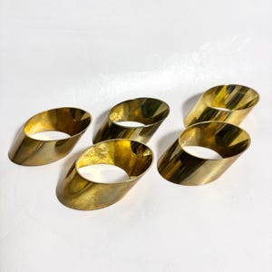 May include: A set of five polished gold-tone metal napkin rings, each featuring a smooth, cylindrical shape with slanted, diagonal cut ends. The metallic surface reflects light, showcasing a bright, reflective finish across all five individual pieces.