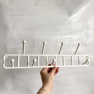 May include: A white wall-mounted coat rack with seven rounded hooks. The rack has a rectangular frame with a grid-like design. The hooks are evenly spaced and extend upwards. The rack is held by a hand against a white background.