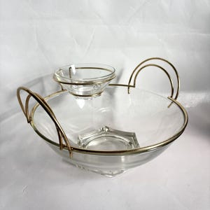 May include: A clear glass serving bowl with a gold-colored metal rim and handles. A smaller glass bowl sits on top. The bowl has a geometric base and a small, built-in compartment.
