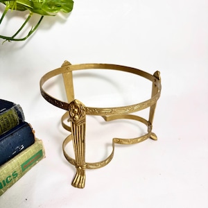 May include: A gold-toned metal stand featuring a circular top ring with an embossed floral pattern, supported by three vertical legs that terminate in claw-style feet. The structure includes a lower heart-shaped frame connecting the three legs.