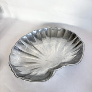 May include: A silver-colored, shell-shaped serving dish. The dish has a scalloped edge and a textured, fan-like pattern radiating from the center. The dish is made of metal and is suitable for serving food or as a decorative item.