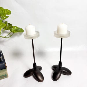 May include: Two tall candle holders featuring thin black metal stems and three-pronged, rounded black bases. Each holder is topped with a circular, frosted glass plate that supports a small, white, cylindrical pillar candle.