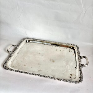 May include: A rectangular silver serving tray with ornate detailing around the edges and two curved handles. The tray has a slightly tarnished surface, showing signs of age and use. The design is classic and elegant.