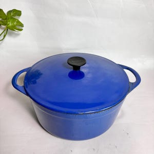 May include: A round, cobalt blue enameled cast iron Dutch oven with two side handles and a matching lid. The lid features a central black knob. The pot has a smooth, glossy finish and a wide, deep basin, providing a sturdy cooking vessel.