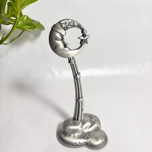 Pode incluir: Uma estatueta de metal prateado com uma lua crescente com um rosto adormecido no topo, ligada por uma haste fina a uma base em forma de nuvem. A base tem gravado o texto "sweet dreams" e pequenas estrelas espalhadas pela haste.