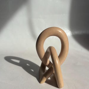 May include: A light beige wooden sculpture with a circular top and intertwined base. The sculpture is smooth and has a natural wood grain appearance. The piece casts a long shadow on a white surface.