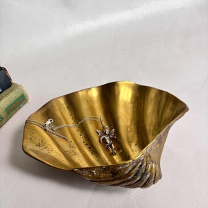 May include: A brass shell-shaped dish with a polished interior and a textured exterior, containing a silver-toned metal necklace featuring a small winged cherub pendant resting inside the bowl of the shell.