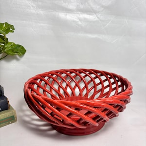 May include: A bright red ceramic bowl featuring a decorative lattice weave design. The bowl has a circular base and a wide, open top with a glossy finish. The interwoven structure creates a series of diamond-shaped openings around the sides.