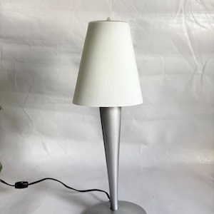 May include: A modern table lamp with a white lampshade and a silver-colored base and stem. The lamp has a conical shade and a round base. A black power cord extends from the base.